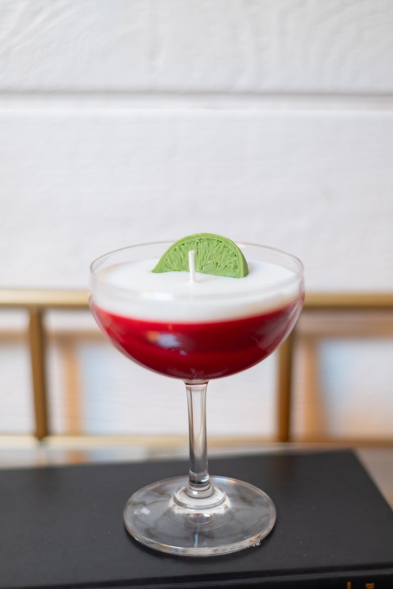 May include: A cocktail-shaped candle with a clear glass, featuring a red base, a white top, and a green lime wedge. The candle has a wick and is designed to resemble a drink.