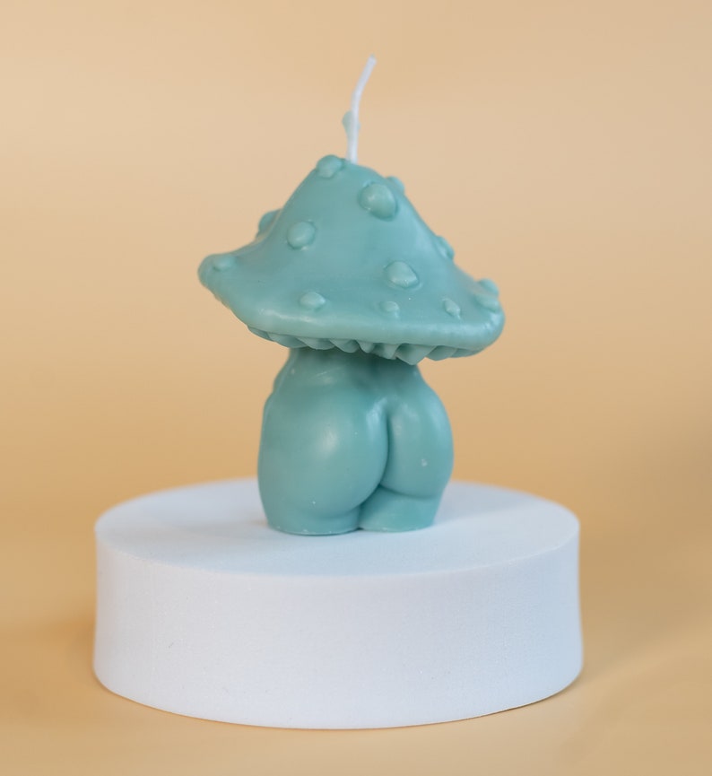 Op de afbeelding: Een teal paddenstoelvormige kaars met een getextureerde dop en een afgeronde basis, die op een figuur lijkt. De kaars staat op een witte, cilindrische sokkel tegen een lichtoranje achtergrond.
