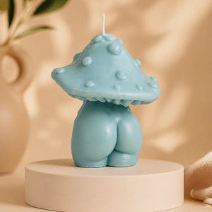 Op de afbeelding: Een lichtblauwe paddenstoelvormige kaars met een getextureerde dop en een afgeronde basis. De kaars wordt gepresenteerd op een kleine, ronde, beige sokkel. De kaars heeft een grillig, artistiek ontwerp.