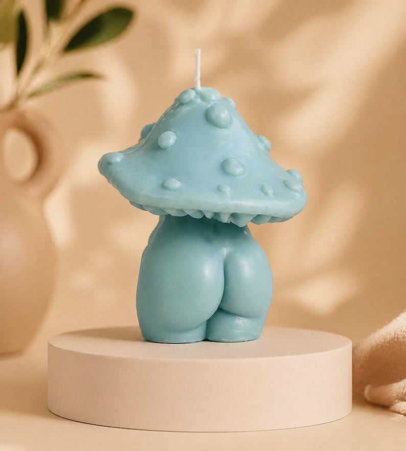 Op de afbeelding: Een lichtblauwe paddenstoelvormige kaars met een getextureerde dop en een afgeronde basis. De kaars wordt gepresenteerd op een kleine, ronde, beige sokkel. De kaars heeft een grillig, artistiek ontwerp.