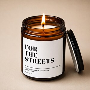 Peut inclure: Une bougie allumée dans un pot en verre ambré avec un couvercle en métal noir. L'étiquette blanche porte le texte noir FOR THE STREETS, NATURAL COCONUT WAX, COTTON WICK, MADE IN CANADA, 283g | Candle Lane. La bougie a une mèche en coton allumée.