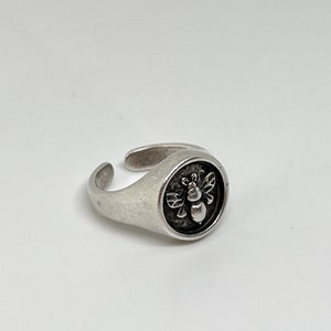 Può includere: Un anello d'argento con un designo a forma di ape. L'ape è al centro dell'anello ed è circondata da un cerchio.