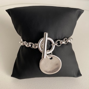 Peut inclure: Un bracelet en argent avec un grand charme rond et un fermoir à bascule. Le bracelet est sur un coussin de velours noir.
