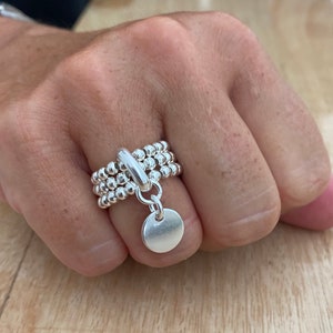 Peut inclure: Une bague en argent avec trois bandes de petites perles et un charme circulaire suspendu. La bague est sur le doigt d'une personne.