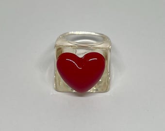 Anel grande de resina com fundo transparente e um coração vermelho.