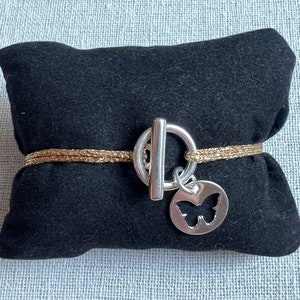 Peut inclure: Un bracelet doré avec un fermoir à bascule argenté et un charm papillon. Le bracelet est présenté sur un coussin en velours noir, mettant en valeur son design délicat et ses détails complexes. Le charm papillon ajoute une touche de fantaisie.