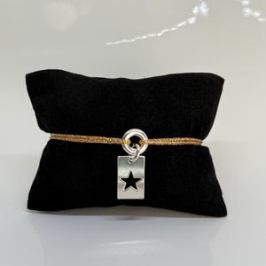 Peut inclure: Un bracelet argenté avec un charm rectangulaire en forme d'étoile. Le bracelet a un cordon doré et est présenté sur un coussin de velours noir. Le bracelet est un bijou simple et élégant.