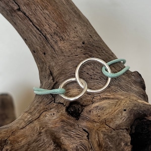 Può includere: Un bracciale in argento con due cerchi interconnessi, collegati da un cordino verde chiaro. Il gioiello è presentato su un pezzo di legno stagionato, evidenziando il suo design minimalista e il contrasto cromatico.