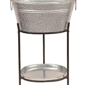Puede incluir: Una tina de metal galvanizado con textura martillada, sobre un soporte de hierro forjado negro. El soporte tiene un estante inferior y patas curvas. La tina tiene dos asas.