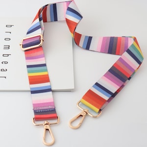 May include: A colorful striped fabric bag strap with gold metal clasps. The strap is made of a woven fabric with a rainbow pattern.