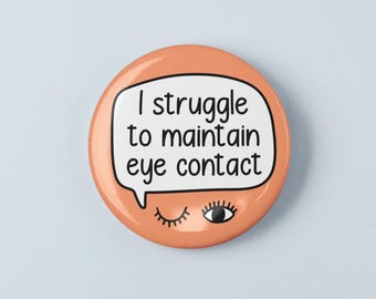 May include: A round peach-colored pinback button featuring a white speech bubble with the black text 'I struggle to maintain eye contact'. Below the speech bubble, there is a graphic of one winking eye and one open eye with black eyelashes.