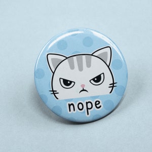 Può includere: Un bottone blu e bianco con un viso di gatto dei cartoni animati e la parola "nope" stampata su di esso. Il gatto ha un'espressione scontrosa con le sopracciglia aggrottate e una smorfia.