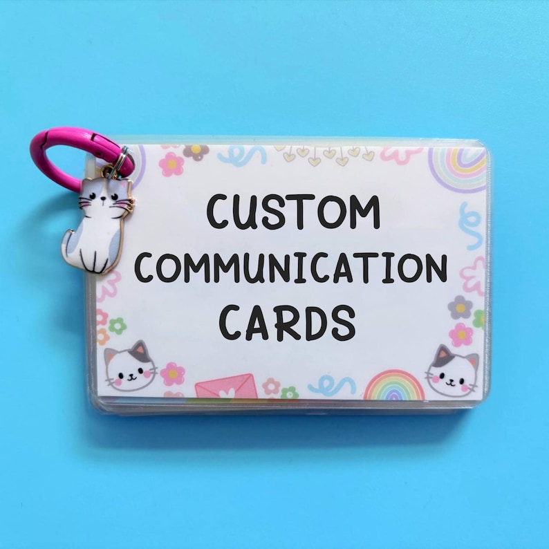May include: A set of custom communication cards in a clear plastic holder, with a pink clasp and a cat charm. The cards have a white background with floral and rainbow designs, and the text "CUSTOM COMMUNICATION CARDS".