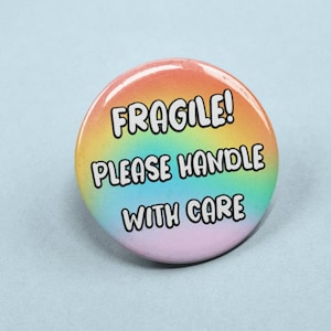 May include: A round button with a rainbow gradient background. The button has the words "FRAGILE! PLEASE HANDLE WITH CARE" in bold, black text. The button is on a light blue background.