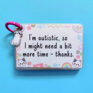 May include: A clear plastic card with the text "I'm autistic, so I might need a bit more time - thanks." is attached to a pink clasp with a cat charm. The card has a floral and rainbow design with cat illustrations.