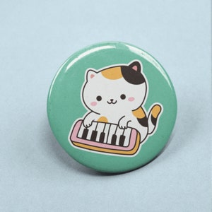 Peut inclure: Un bouton vert avec un mignon chat de dessin animé jouant d'un clavier rose et jaune. Le chat a une fourrure blanche, des taches orange et noires et un nez rose.