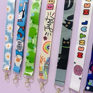 May include: Six fabric lanyards with metal lobster clasps. Designs include white daisies on blue, a goose with the text 'TODAY I AM A SERIOUS GOOSE', green frogs, a rainbow with 'BE KIND', black cats on grey, and 'MENTAL HEALTH' with a brain icon.