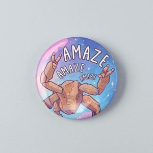May include: A circular pinback button featuring a tan, rocky, multi-limbed creature making peace signs with its hands against a blue and purple galaxy background. The text "AMAZE" is repeated three times in a white, hand-drawn font above the creature.