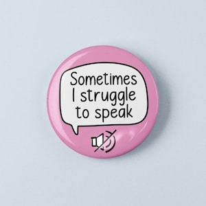 May include: A circular pink pinback button featuring a white speech bubble with the black text "Sometimes I struggle to speak" and a black icon of a speaker with a diagonal line through it, indicating a muted or silent audio status.