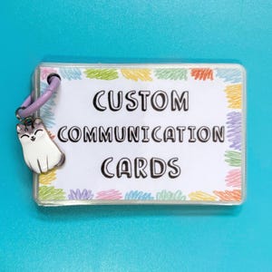 May include: A set of rectangular communication cards held together by a light purple ring. The cards feature the text CUSTOM COMMUNICATION CARDS in black, surrounded by a border of colourful scribbles. A small white cat charm hangs from the ring.