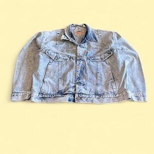 May include: Acid wash denim jacket with two chest pockets and two side pockets. The jacket has a button-up front and a collared neckline. The denim is a light blue color with a faded, distressed look. The jacket is laid flat on a yellow background.