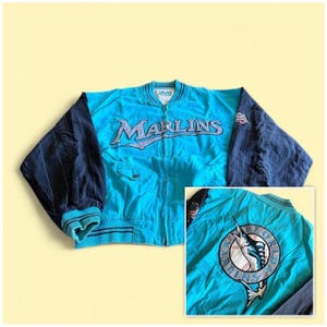 May include: A turquoise and dark blue jacket featuring the word MARLINS embroidered on the front. The sleeves are dark blue with thin pinstripes. The back displays a large circular patch with a marlin fish graphic over a baseball design.