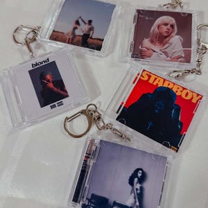 Könnte beinhalten: Klare Schlüsselanhänger aus Kunststoff in Form von CD-Hüllen mit Albumcovern verschiedener Künstler. Die Schlüsselanhänger haben silberne Metall-Schlüsselringe. Die Albumcover zeigen Bilder von Personen, Text und Albumcover.