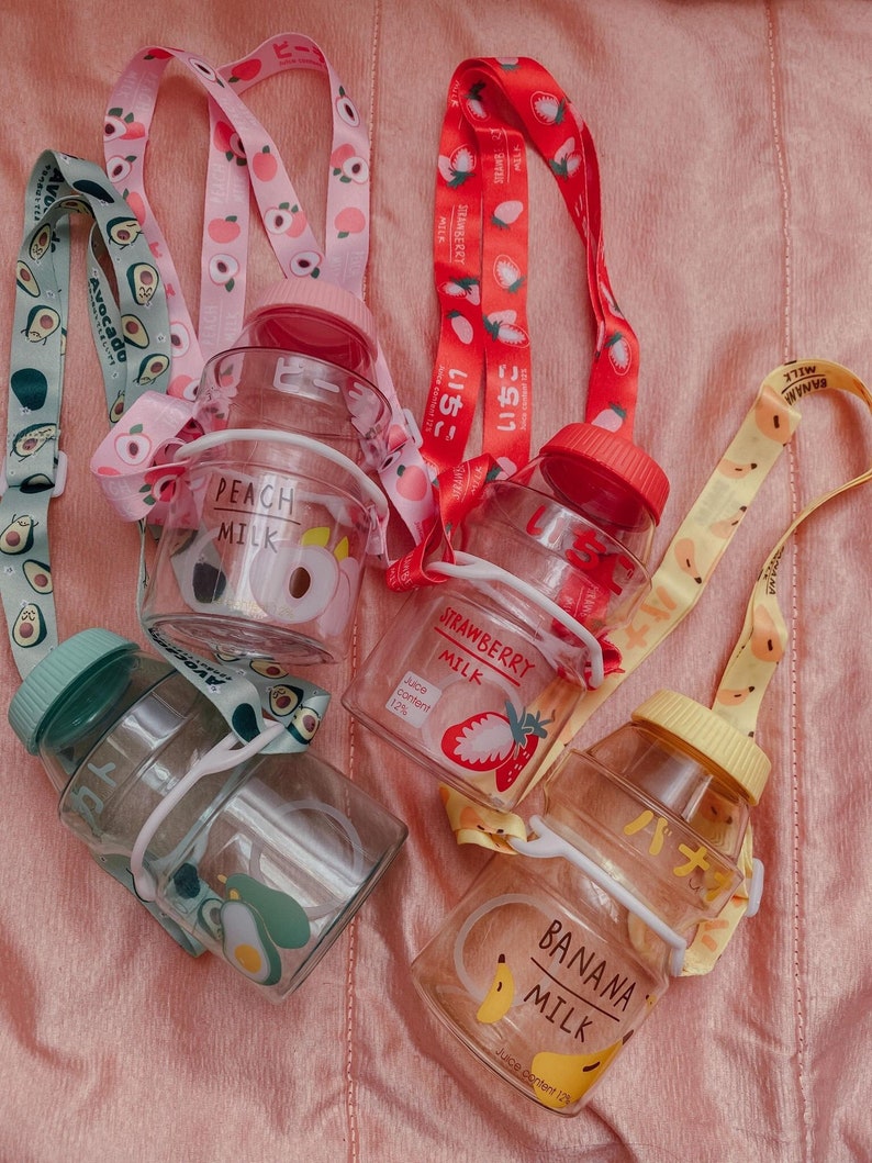 May include: Four clear plastic water bottles with colorful straps. Each bottle has a different fruit design and text: avocado, peach, strawberry, and banana. The text on the bottles reads "Avocado Milk", "Peach Milk", "Strawberry Milk", and "Banana Milk".