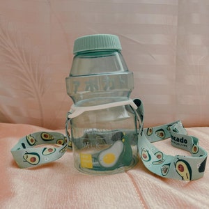 May include: A light green plastic water bottle with a light green lid and a white strap. The strap has a pattern of green avocados with faces on them. The bottle has the word "MILK" printed on it and the text "Avocado content 100%" printed on a yellow sticker.