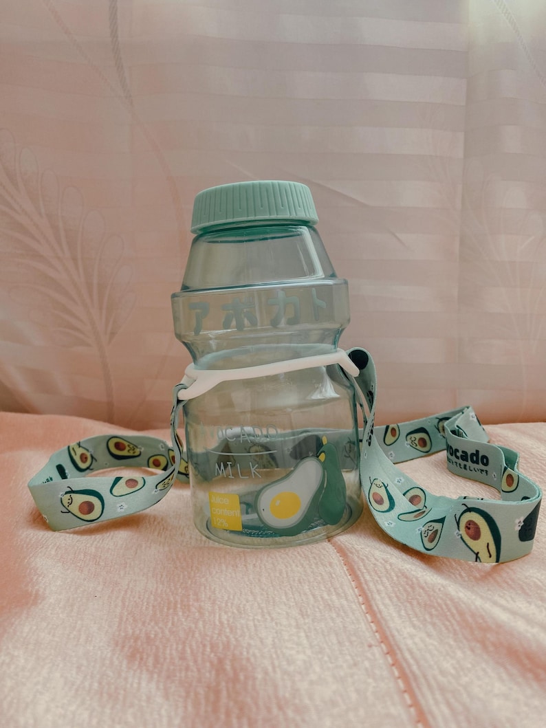 May include: A light green plastic water bottle with a light green lid and a white strap. The strap has a pattern of green avocados with faces on them. The bottle has the word "MILK" printed on it and the text "Avocado content 100%" printed on a yellow sticker.