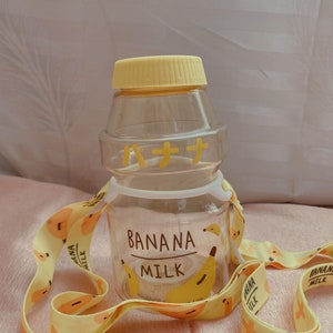 May include: A clear plastic water bottle with a yellow lid and a yellow strap. The bottle has a banana design and the text "Banana Milk" on it. The strap has a repeating banana design.