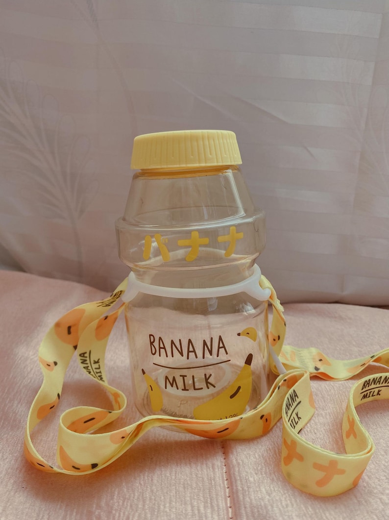 May include: A clear plastic water bottle with a yellow lid and a yellow strap. The bottle has a banana design and the text "Banana Milk" on it. The strap has a repeating banana design.