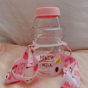 May include: A pink and clear plastic water bottle with a peach design and the text "PEACH MILK" on the front. The bottle has a pink strap with peach designs.