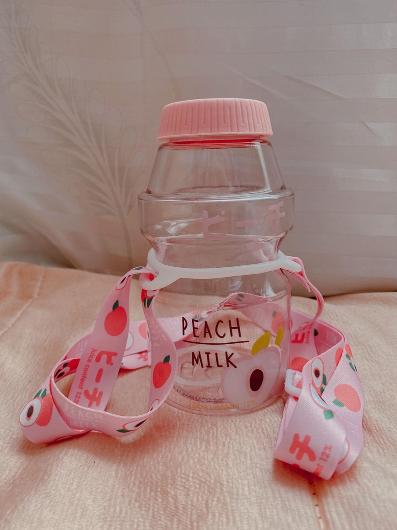 May include: A pink and clear plastic water bottle with a peach design and the text "PEACH MILK" on the front. The bottle has a pink strap with peach designs.