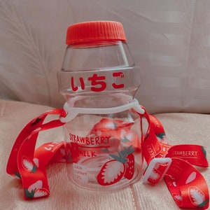 May include: A clear plastic water bottle with a red lid and a red strap. The bottle has a strawberry design and the text "STRAWBERRY MILK" and "Juice content 12%". The bottle also has the Japanese text "いちご" on it.