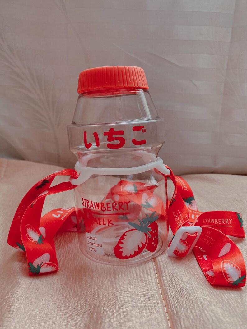 May include: A clear plastic water bottle with a red lid and a red strap. The bottle has a strawberry design and the text "STRAWBERRY MILK" and "Juice content 12%". The bottle also has the Japanese text "いちご" on it.