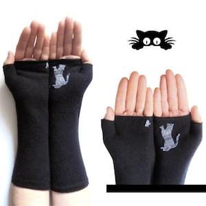 Puede incluir: Guantes sin dedos negros con un diseño de silueta de gato gris. Los guantes están hechos de un material suave y están diseñados para cubrir la mano hasta los nudillos, dejando los dedos libres. También se ve una cara de gato negro.