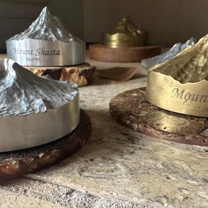 May include: Metal mountain sculptures, including Mount Shasta and Mount Hood, sit on decorative coasters. The sculptures are silver and gold-toned, with engraved text. The Mount Shasta sculpture is 14,179 ft. high. The Mount Hood sculpture is 11,249 ft. high.