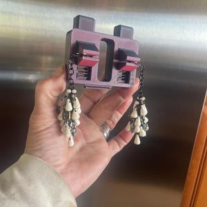 May include: A pair of white beaded dangle earrings with black chains hanging from a 3D printed black and red earring holder.