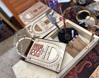 May include: A selection of wooden jewellery-making tools and accessories. The image shows a wooden box containing a caliper, wire coils, and a wooden tool with numbered markings. The tools are arranged on a wooden surface.
