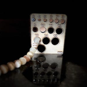 May include: A wooden bead sizer tool with multiple holes of varying sizes, labeled with numbers. A strand of light-colored beads is partially inserted into one of the holes. The tool is reflected in a dark, glossy surface.