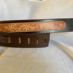 May include: A brown leather guitar strap with a tan leather strip featuring a leaf pattern. The strap has five adjustment holes.