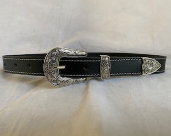 May include: Black leather belt with white stitching and a silver-coloured buckle. The buckle and belt tip feature floral and scrollwork designs. The belt is approximately 3.8 cm wide.