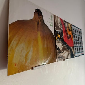 May include: Three vinyl records displayed on a white wall using record holders. The first record features a large, close-up image of a yellow onion. The second record has a colourful abstract design with the text "JOGUAB" on the bottom. The third record has a black and white photo of a group of people.