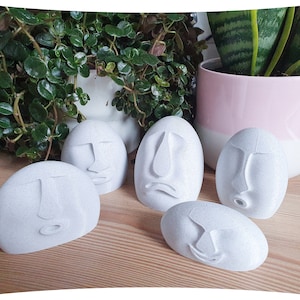 May include: Five white, round, smooth, decorative objects with different facial expressions. Each object has a unique face carved into the surface. The objects are arranged on a wooden surface.