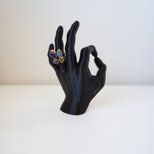 May include: A black hand-shaped jewelry display stand holding a gold ring with blue and brown gemstones. The ring has a cluster of teardrop and oval-shaped stones. The display stand is on a white surface.