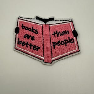 Peut inclure: Écusson brodé en forme de livre ouvert, avec des pages roses et une bordure blanche. Le texte "books are better than people" est brodé en noir sur les pages. L'écusson a des accents noirs sur le dos et les coins.