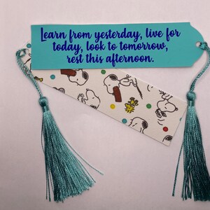 Puede incluir: Un marcap&aacute;ginas hecho a mano con una borla azul claro. El marcap&aacute;ginas presenta un fondo blanco con personajes de Snoopy y el texto "Learn from yesterday, live for today, look to tomorrow, rest this afternoon."