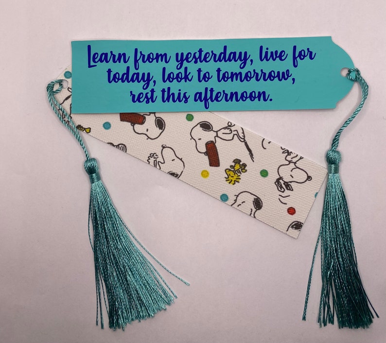 Puede incluir: Un marcap&aacute;ginas hecho a mano con una borla azul claro. El marcap&aacute;ginas presenta un fondo blanco con personajes de Snoopy y el texto "Learn from yesterday, live for today, look to tomorrow, rest this afternoon."