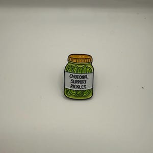 May include: A white ceramic mug with a black handle and the text "But First, Coffee" in a bold, black font. The mug is set against a blurred background. The mug appears to be approximately 10 cm tall.